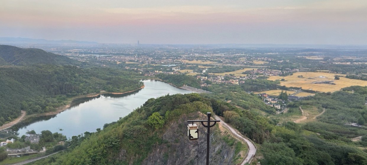 Photograph from the ridge above 康门水库 looking down the water and out over the plain beyond it, with the walkway's lamp post in the foreground on the rock