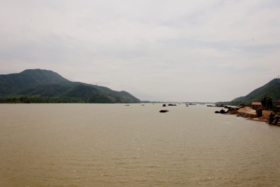 Wide brown river with barges and dredging plant on a low bank, hills at either side